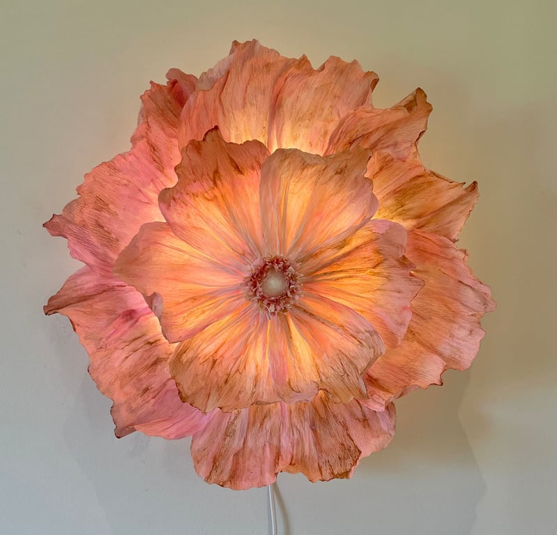 Lyserød og orange blomsterlampe med bløde, bølgede kronblade, der udsender et varmt, diffust lys mod en lys væg.