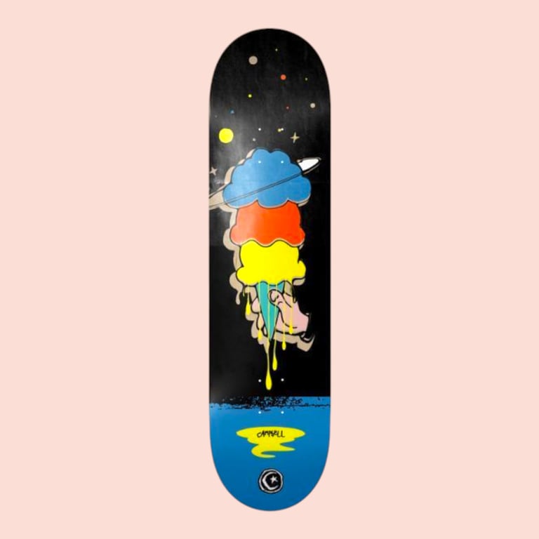 Sort skateboard med illustration af en hånd, der holder en smeltende, flerfarvet is med blå, rød og gul kugle, omgivet af stjerner og en planet med ringe på en nattehimmel.