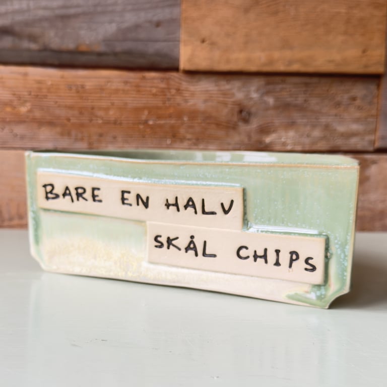 Keramisk chips-skål med tekst på dansk "Bare en halv skål chips" foran trævæg.