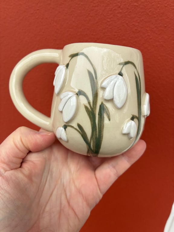 Hånd, der holder en cremefarvet keramikkrus med reliefferede og malede hvidt sneprydningsblomster mod en rød baggrund.