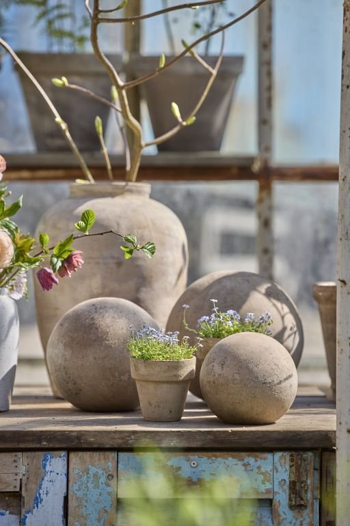 Stilleben med lerkrukker, kugler og små blomster i potter på et træbord foran et vindue.