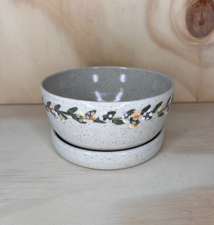 Keramisk skål med låg, dekoreret med malede blomster og blade, placeret på lyst træbord.