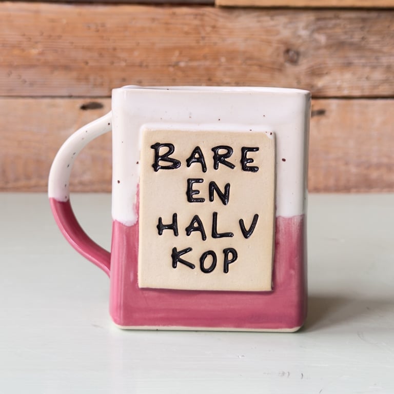 Keramikkop med rektangulært design, lyserød og hvid glasur, tekst "Bare en halv kop" på forsiden.