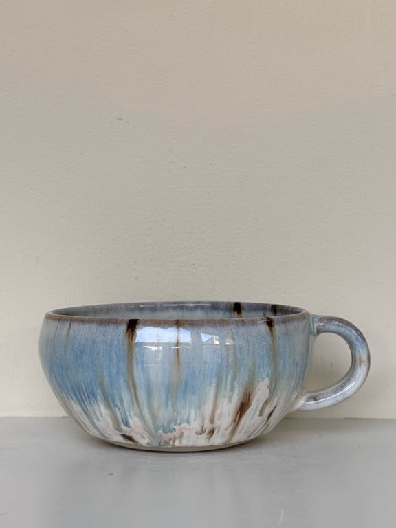 Blå og brun keramikskål med hank mod beige væg.