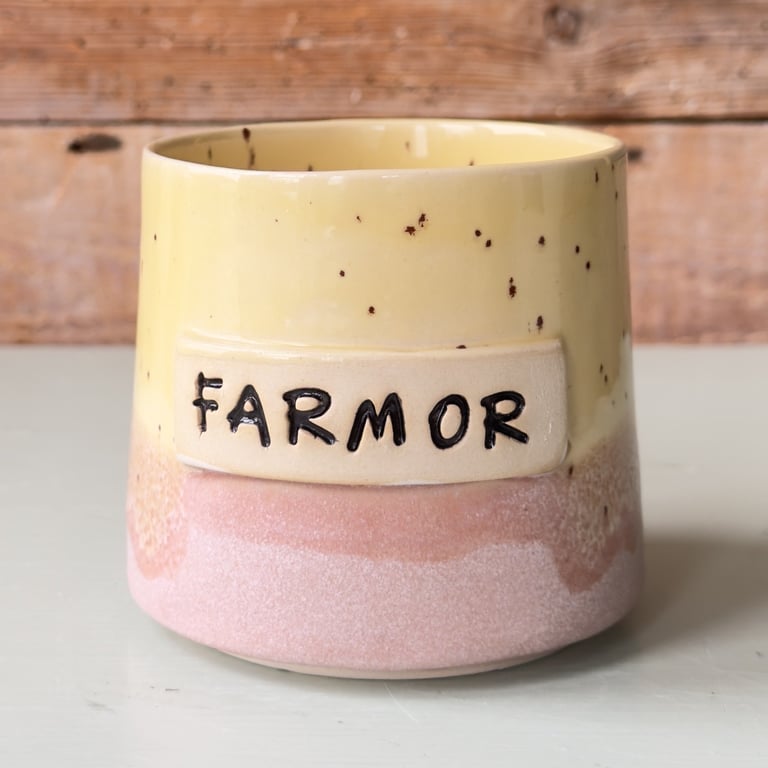 Keramikkrus med teksten "Farmor" i sort på beige label, kruset er gult øverst og lyserødt nederst.