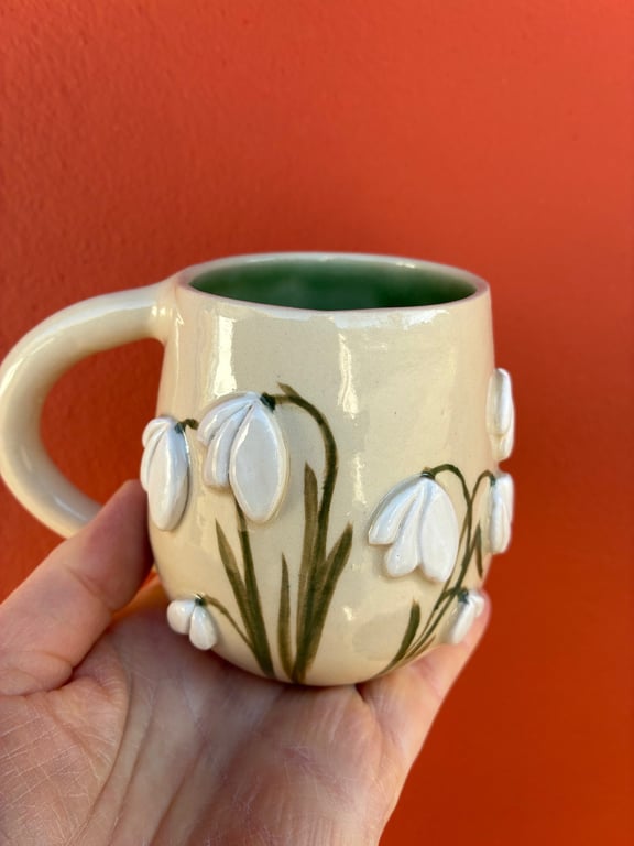 En hånd holder en beige keramikkop med grøn inderside og reliefdekoration af hvide vintergækker mod en orange væg.