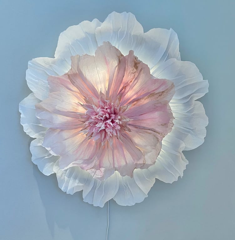 Dekorativ væglampe formet som en stor blomst med lyserøde og hvide lag af blomsterblade.