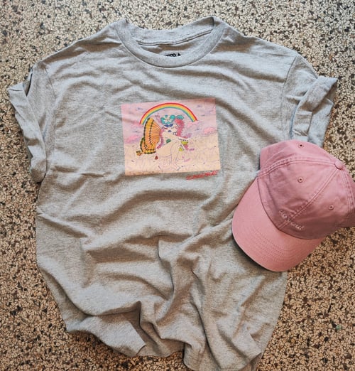 Grå T-shirt med farverigt motiv af en enhjørning og regnbue, lagt fladt på mønstret gulv med lyserød kasket ved siden af. (2)