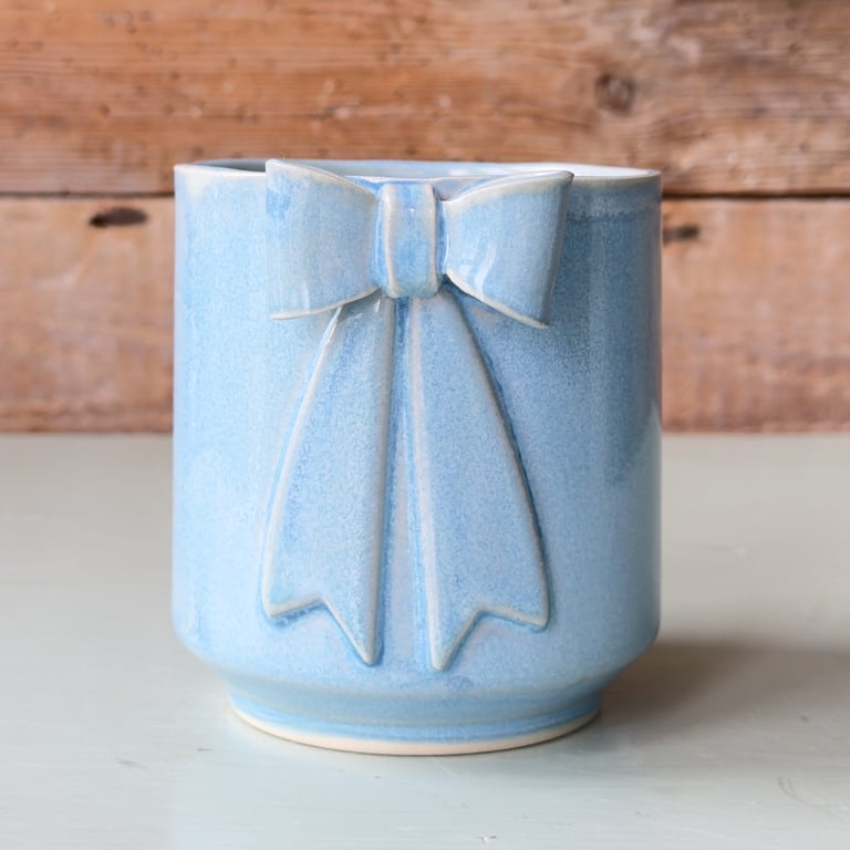 Blå keramikkrukke med reliefformet sløjfe foran, placeret på en lys overflade med en træbaggrund.
