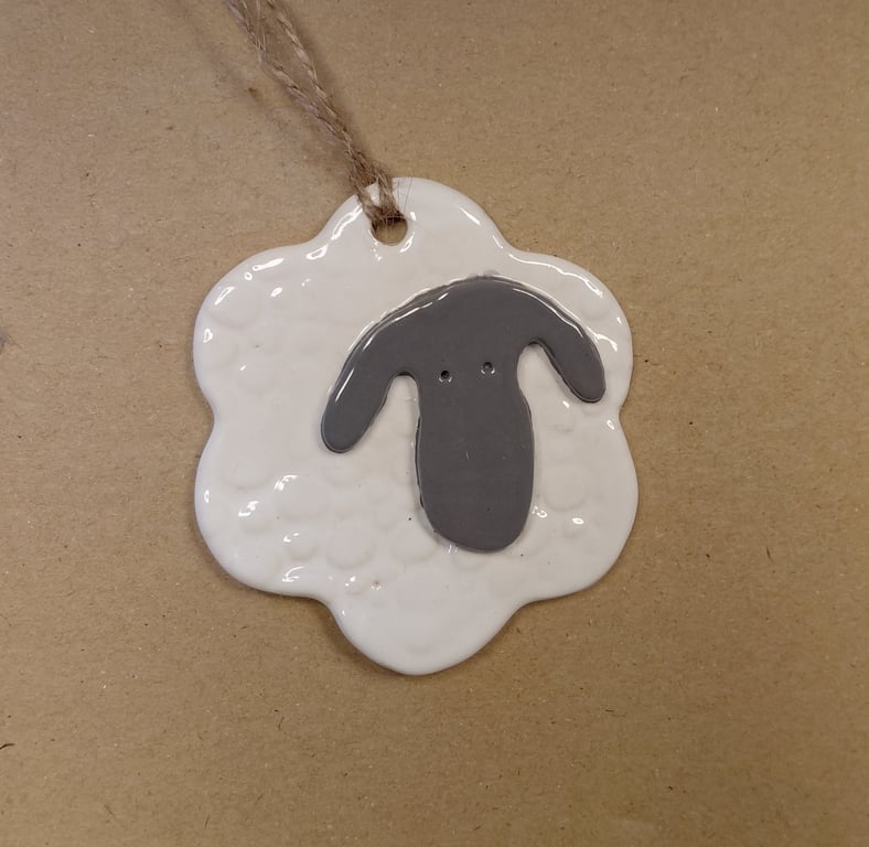 Hvid keramisk juleophæng formet som et får med gråt ansigt og ører, hængende i en grov snor mod en brun baggrund.