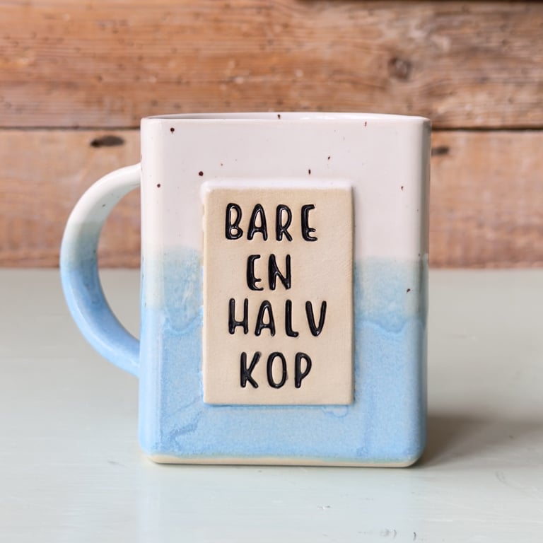 Firkantet keramisk krus med blå og hvid glasur og teksten "Bare en halv kop" på forsiden.