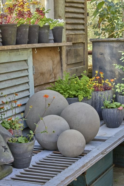 Billede af en havebænk med forskellige grå urtepotter og store dekorative beton-kugler, omgivet af grønne planter og blomster i flere potter.