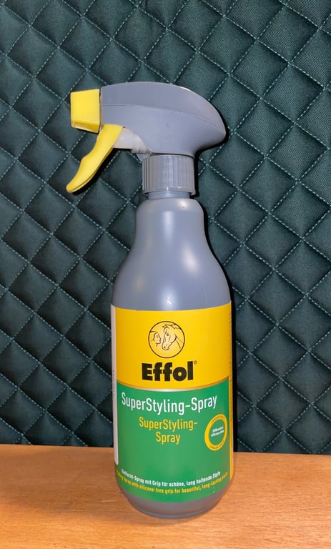 Sprayflaske med gul top og etiket med teksten "Effol SuperStyling-Spray" på grøn og gul baggrund, står på et træbord foran en quiltet sort baggrund.