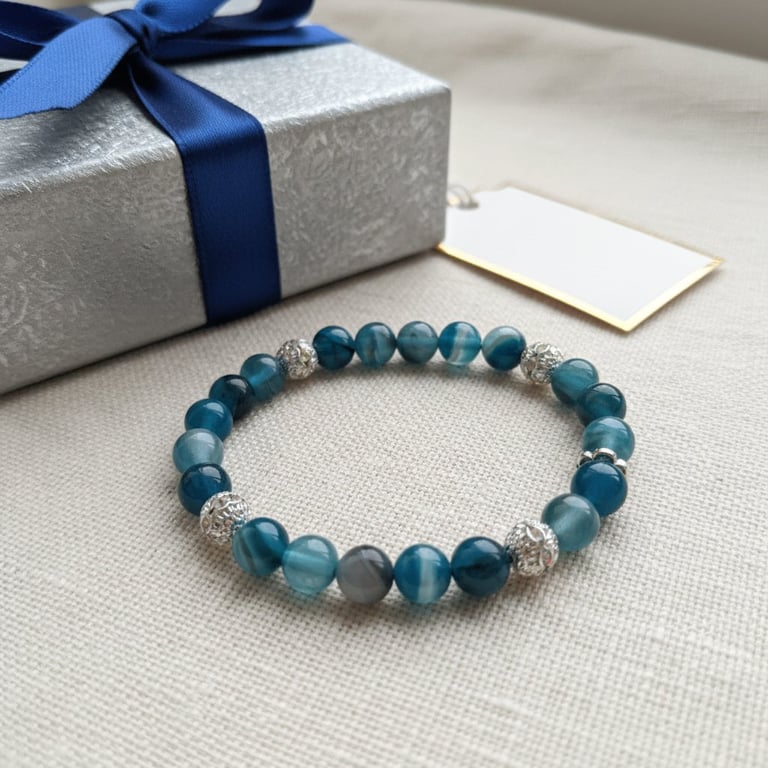 Armbånd af blå og turkise perler med sølvdetaljer liggende på en beige overflade ved siden af en sølvindpakket gaveæske med blå bånd og et blankt, hvidt kort.