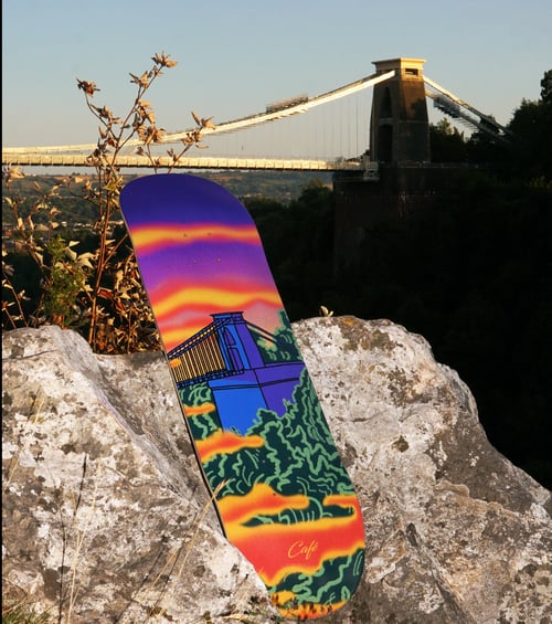 Skateboard med farverigt design af en bro, der står op ad en stor sten med en rigtig bro i baggrunden ved solnedgang. (1)