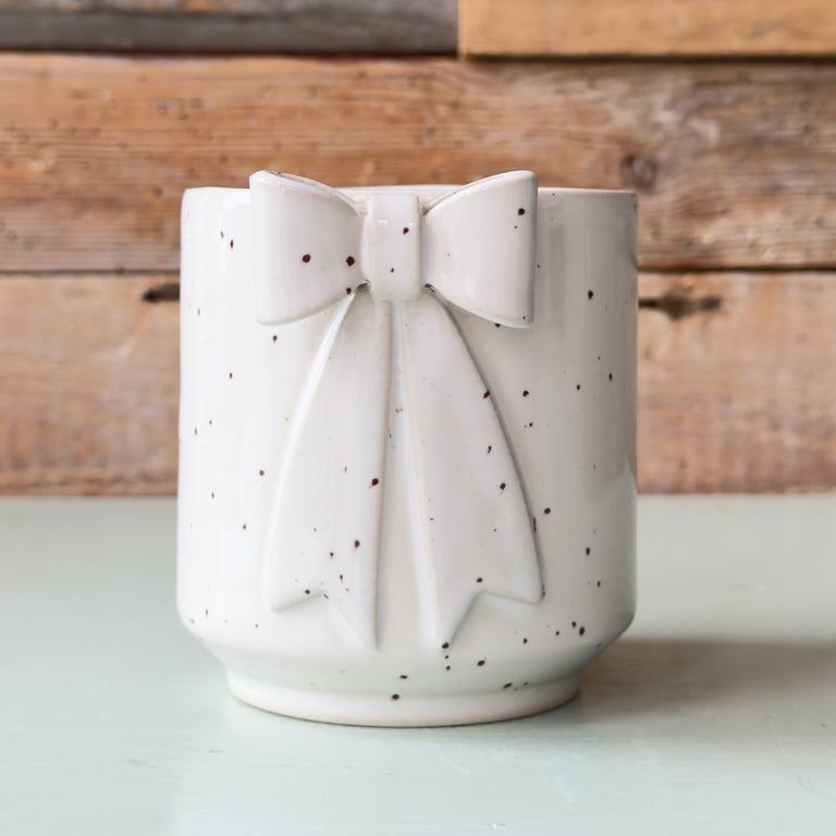 Hvid keramisk kop med dekorativ sløjfe og små brune pletter, placeret på en lys flade med træbaggrund.