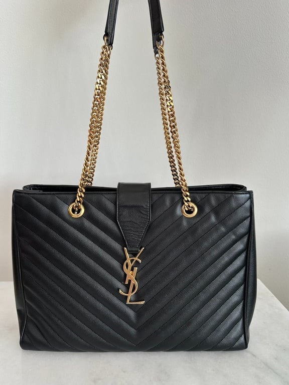 Sort Yves Saint Laurent skuldertaske i skind med gulddetaljer og kæderem, placeret på et lyst marmorbord.