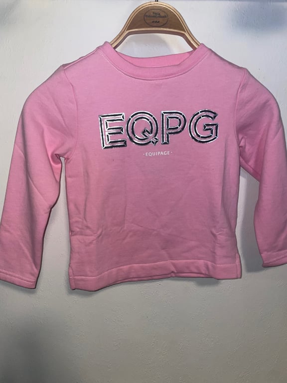 Lyserød langærmet børnetrøje med teksten "EQPG" og "EQUIPAGE" på forsiden, hængende på en træbøjle.