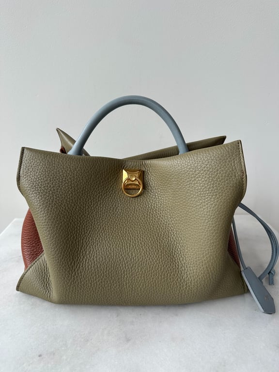 Taske i grågrøn læder med blåt håndtag og guldfarvet lås, placeret på hvid overflade foran hvid væg.