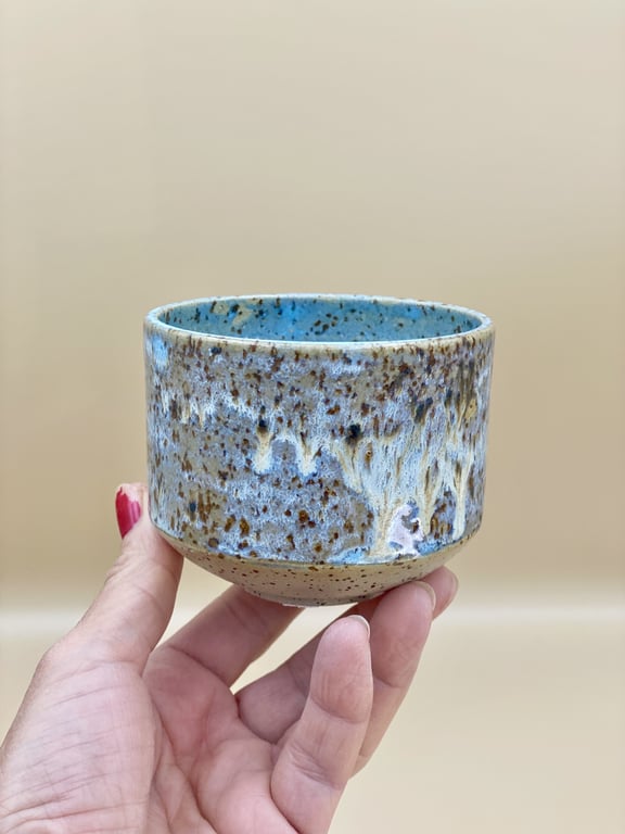 Hånd, der holder en lille keramisk kop med blå og brune glasurer og en spættet tekstur mod en neutral beige baggrund.
