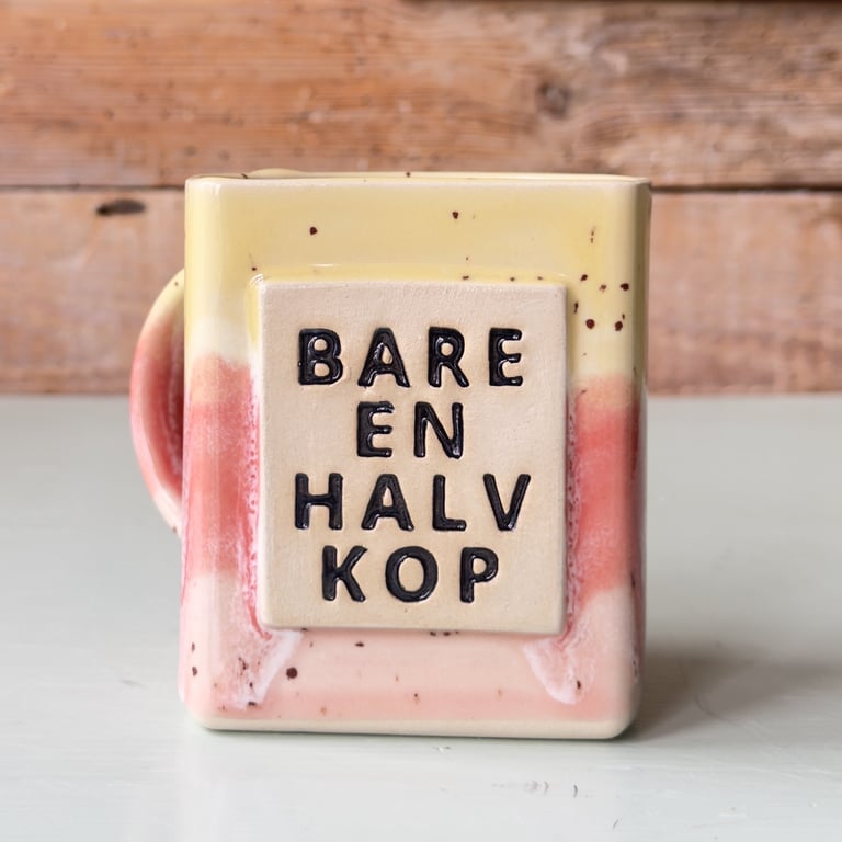 Ceramic krus med tekst "BARE EN HALV KOP" i sort skrift på beige firkant, med gul, rosa og cremefarvet glaseret baggrund.
