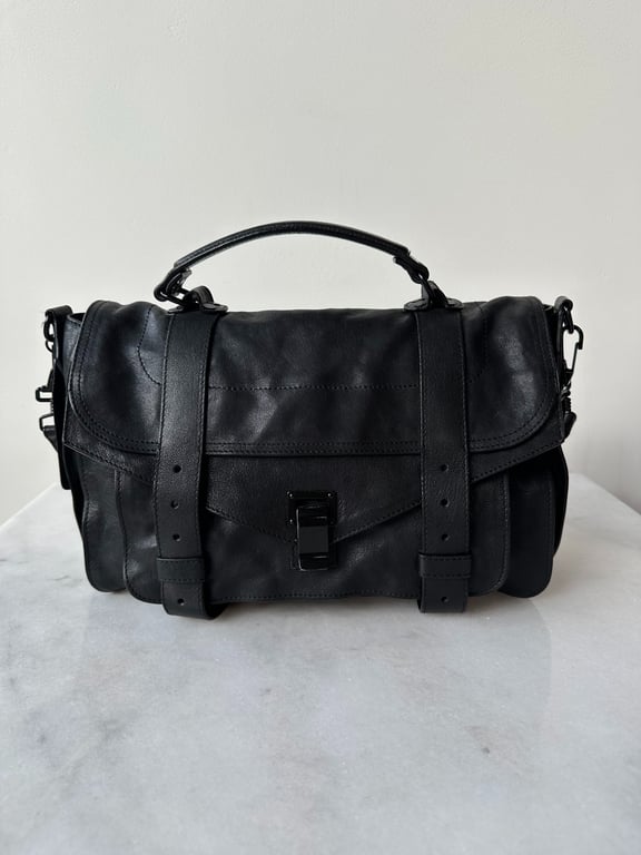 Sort lædertaske med hank og spænder foran, placeret på marmorbord.