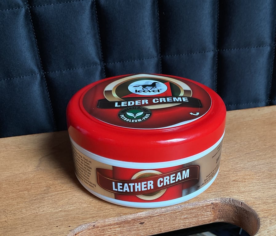 Beholder med rød hætte, mærket "Leder Creme" og "Leather Cream", placeret på et træbord foran en sort polstret baggrund.
