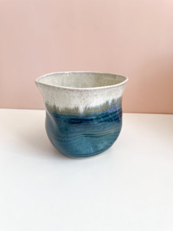 Keramikkop med blå og hvide glasurer stående på hvid overflade mod lyserød baggrund.