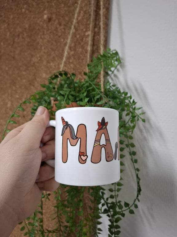 Hånd der holder en hvid kop med bogstaver dekoreret med enhjørning og hjerte, foran en hængende grøn plante.