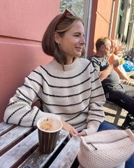 Ung kvinde med brunt hår iført stribet trøje og solbriller i håret sidder ved bord udendørs med en kop kaffe foran sig. (1)