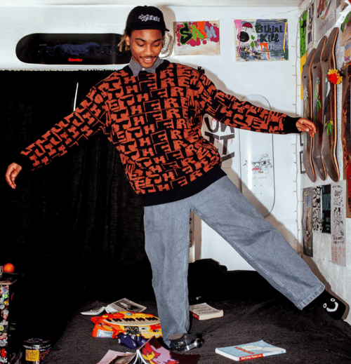 Ung mand i sort kasket og orange-svarthætte trøje balancerer på seng i et værelse med skateboards og plakater på væggene. (2)
