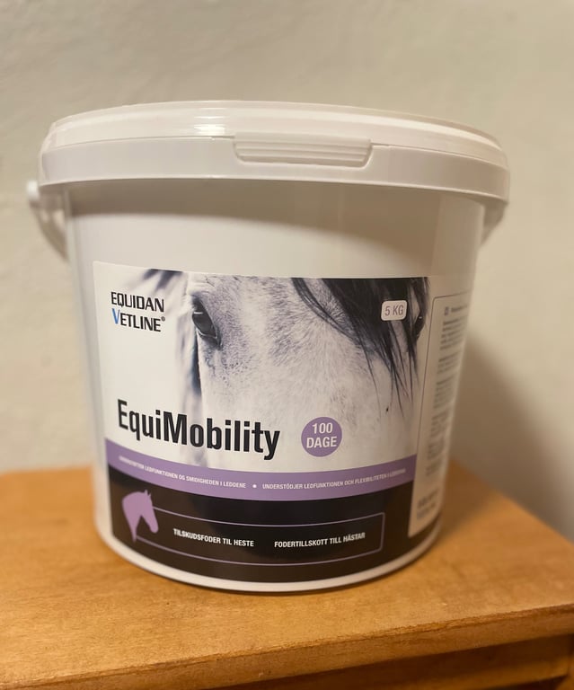Nahbillede af en spand med hestefoderet EquiMobility fra Equidan Vetline, 5 kg, tilskudsfoder til heste, stående på et træbord.