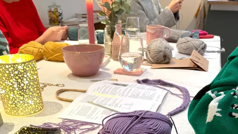 Flere personer sidder ved et bord med strikkegarn, strikkepinde, opskriftsbog, drikkeglas, krus og levende lys.