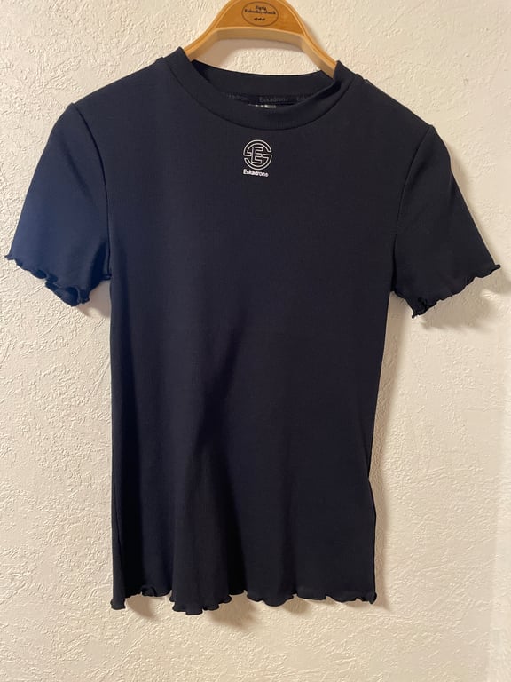 Sort kortærmet t-shirt med bølget kant ved ærmer og bund, hængende på en træbøjle mod en hvid væg.