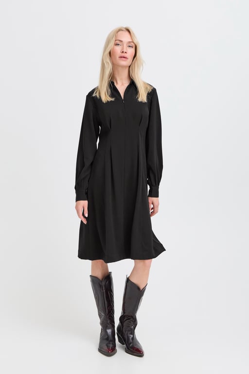 Kvinde iført sort, langærmet midiklæde og mørke cowboystøvler stående mod hvid baggrund.