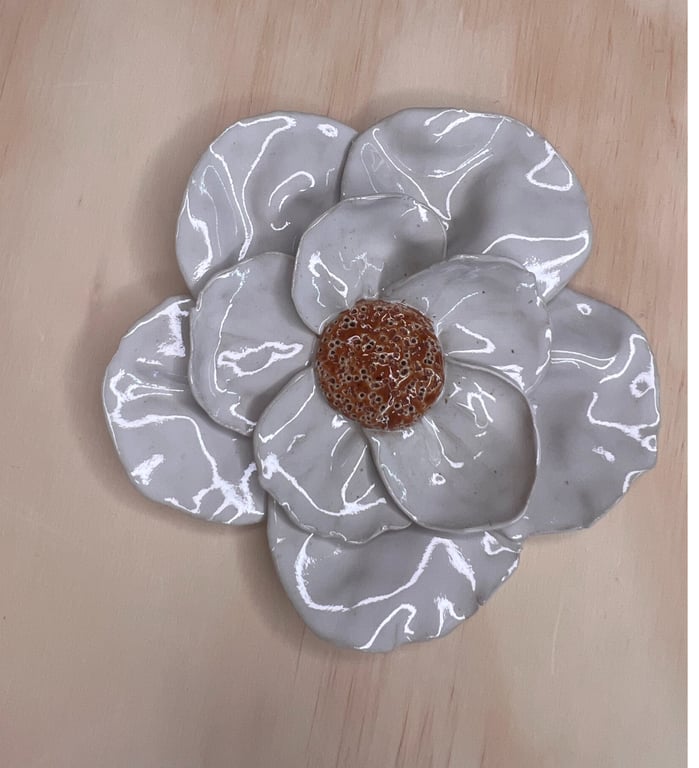 Glaseret keramisk blomst med hvide kronblade og en brun midte, lagt på en trægulvsoverflade.