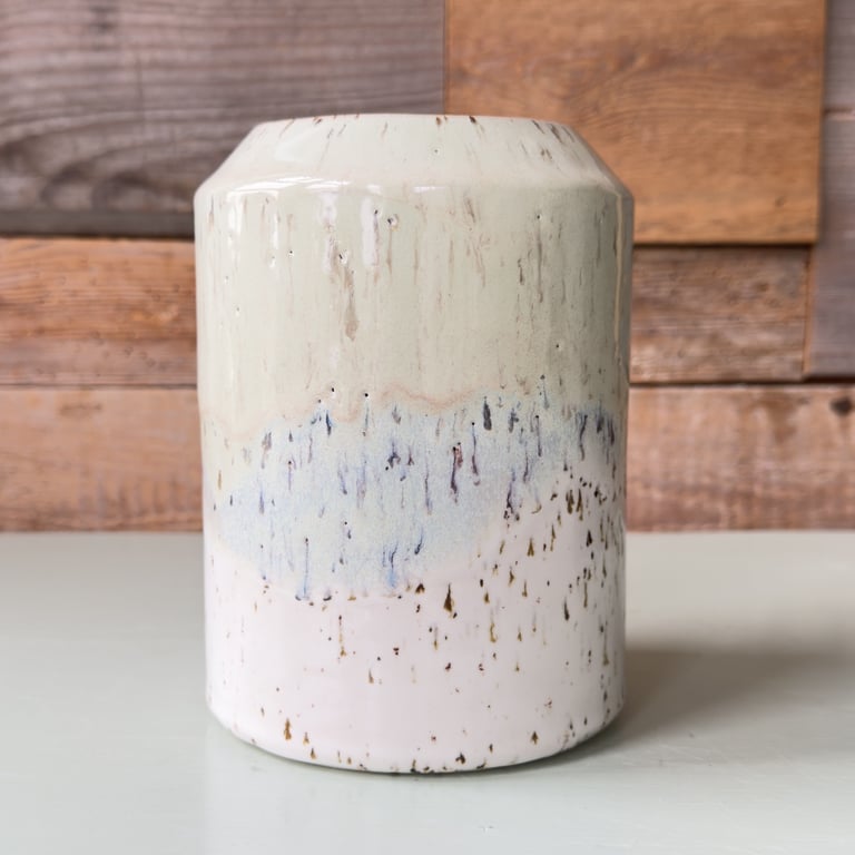 Keramisk vase med beige, blå og hvid glasur, placeret på en grå overflade med en trævæg i baggrunden.