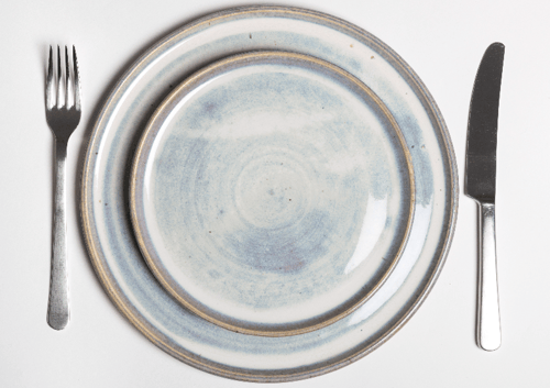 Et tomt spisebord med en blå og cremefarvet tallerken, en kniv og en gaffel på en hvid baggrund.