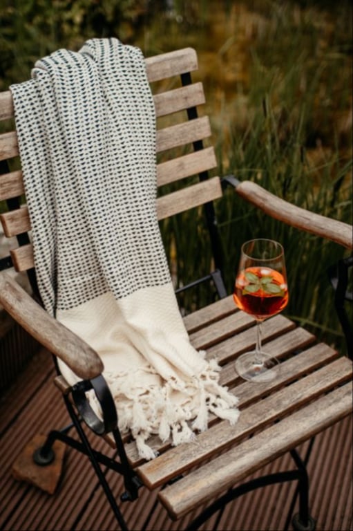 Trækstol i træ med sort metalramme, dækket af et strikket tæppe og et vinglas med orange drink og pynt, placeret udendørs på et trædæk.