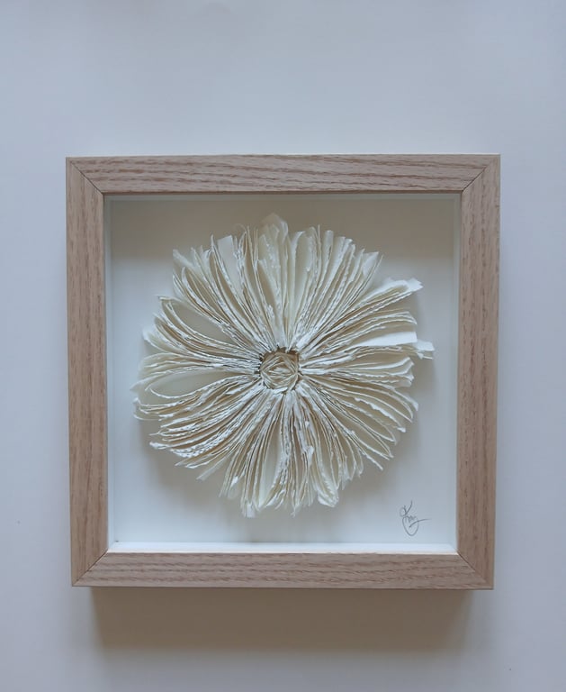 Indrammet kunstværk bestående af en blomst formet af foldede og rullede sider fra en bog med naturfarvede sider mod en hvid baggrund.