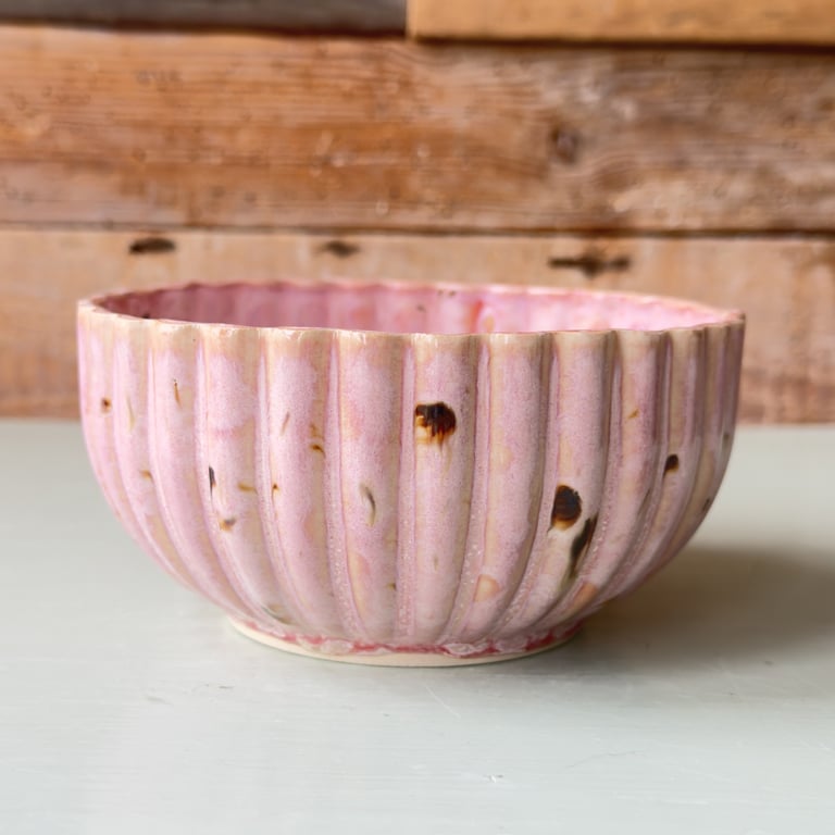 Rund rosa keramisk skål med rillede mønstre og mørke pletter, placeret på hvid overflade med træbaggrund.