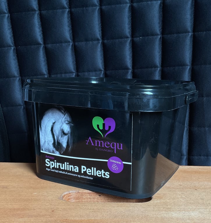 Sort plastbeholder med label til "Amequ Spirulina Pellets" til heste, stående på træbord foran mørk quiltet baggrund.