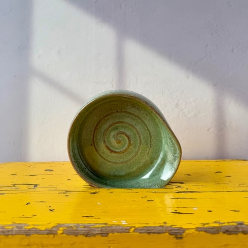 Grøn keramikskål med spiralformet mønster, placeret på et gult, slidt træbord mod en hvid væg med solskygger. (1)
