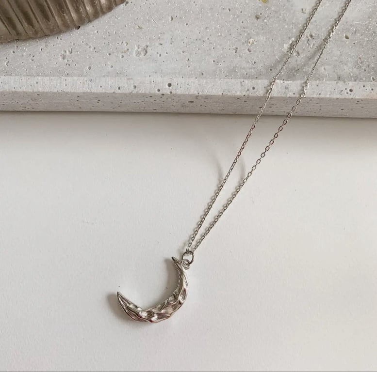 Sølvlignende halvmåneformet vedhæng på en fin kæde lagt på hvid overflade ved siden af en grå, stenagtig genstand. (4)