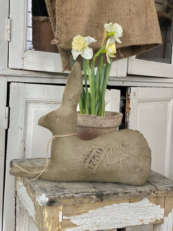 En stofkanin med syninger og teksten "Rjedson 1759" sidder på en rustik, afskallet trækiste foran en lerpotte med hvide og gule påskeliljer.