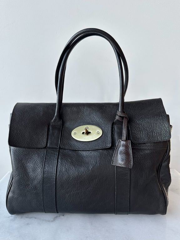 Sort læderhåndtaske med to håndtag og sølvfarvet drejelås på fronten, placeret på marmorbord foran hvid væg.