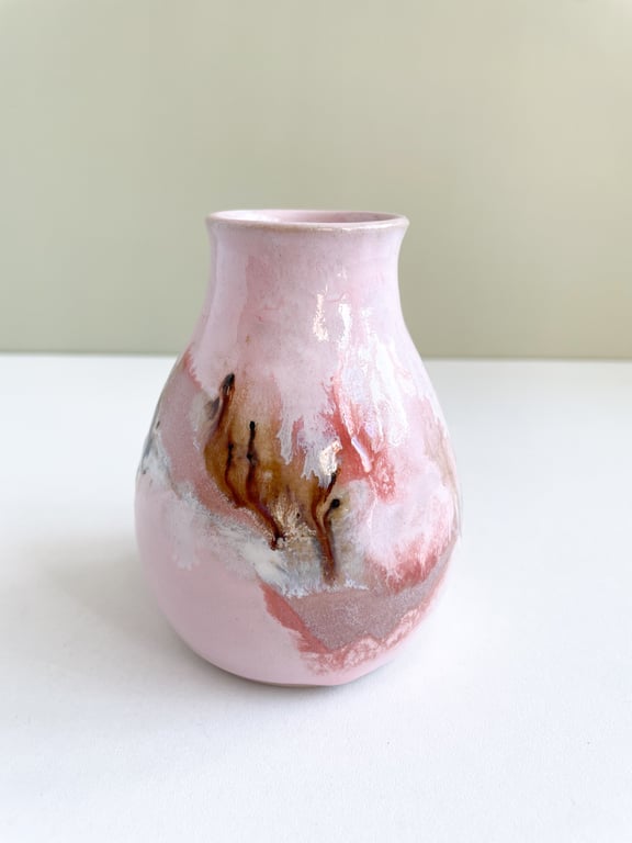 Lyserød keramisk vase med brun, hvid og rød glasurdetaljer, placeret på hvid overflade mod beige væg.