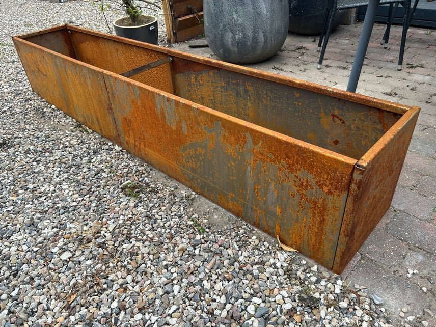 Lang, rusten metal plantekasse placeret på grus ved en terrasse med store grå krukker og havemøbler i baggrunden.