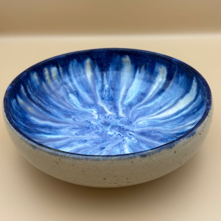Keramisk skål med blå og hvide glasurstriber indvendigt, beige yderside, placeret på lys baggrund.