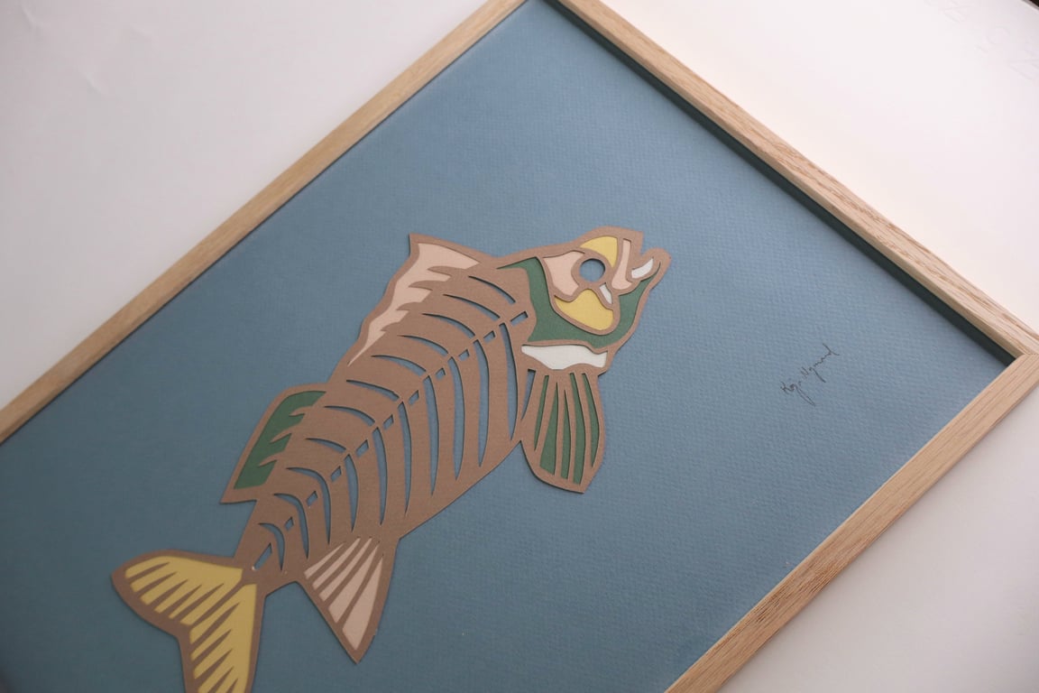 Billedet viser en kunstnerisk tegning af en fisk i brun, grøn og gul på en blå baggrund, indrammet i en lys træramme.